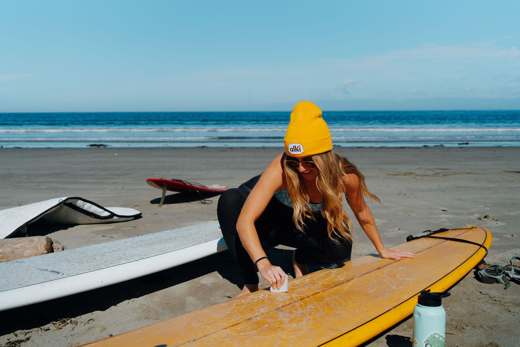 woman wearing yellow beanie waxes a surf board
